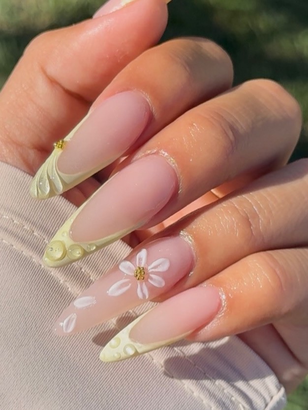 Hand with long almond nails featuring yellow French tips, white daisy nail art and gold accents